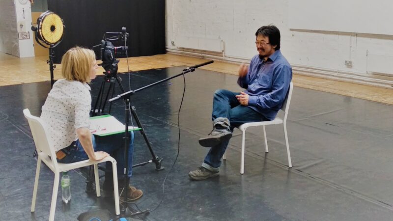A photograph of a man being interviewed in a large space, both the interviewer and the interviewee are sitting on chairs