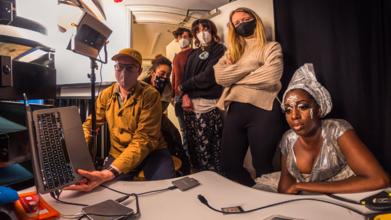 A team of people sit in a small editing studio looking at a monitor. They wear face masks. Electronic equipment