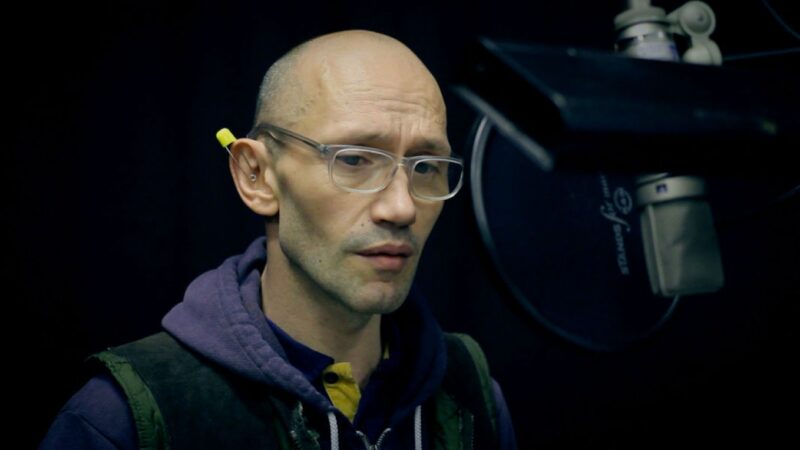 A photograph of a bald man with glasses in front of a microphone