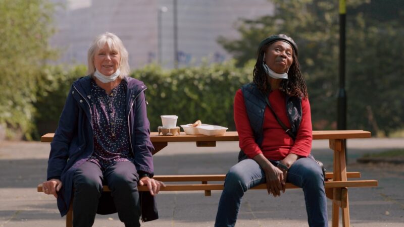 Two women sat on a bench having coffee and food