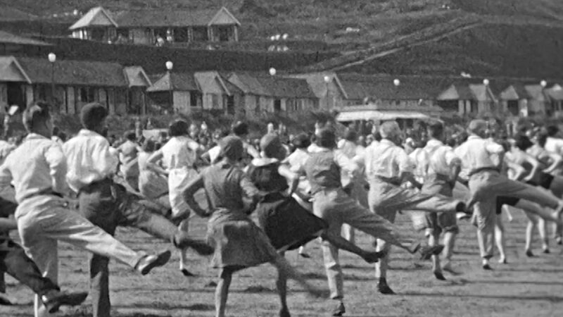An archive black and white image of people exercising on a beach. The men wear white shirts and light trousers and the women wear pinafore's and bonnets