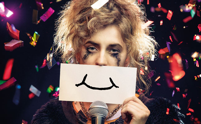 A young woman with messy hair and mascara running down her face standing in front of a mic, holding a drawing of a frown over her mouth.