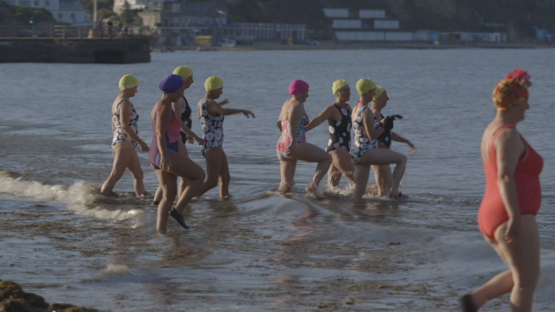 A group of women in colourful swimming costumes and caps, wading into the sea