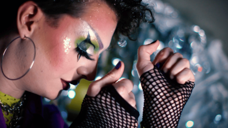 Close-up image of a face and hands. The face features elaborate make-up and a big hoop earring. Fingerless, fishnet gloves are worn on the hands.