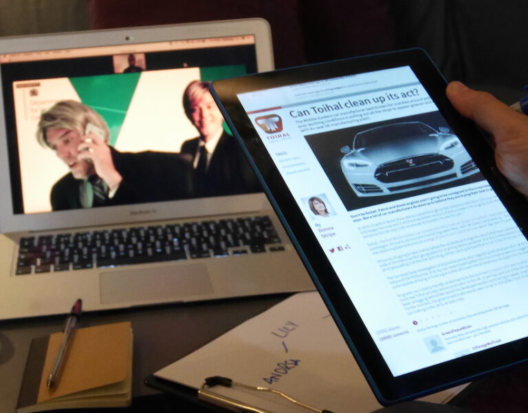 Hand holding tablet showing an article, in front of a laptop playing a video, on a table with a mug of tea and a notebook