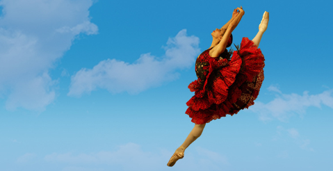 A female ballet dancer in a bright red dress jumps through the air with her arms raised. The background features a blue sky with wispy white clouds.