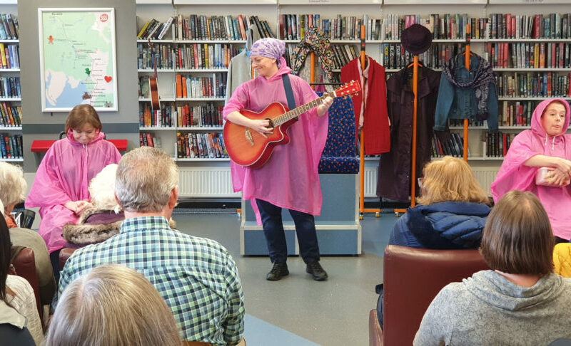 A group of people in a library.One person stands, playing a guitar, wearing a pink raincoat. Some of the seated audience also wear pink raincoats