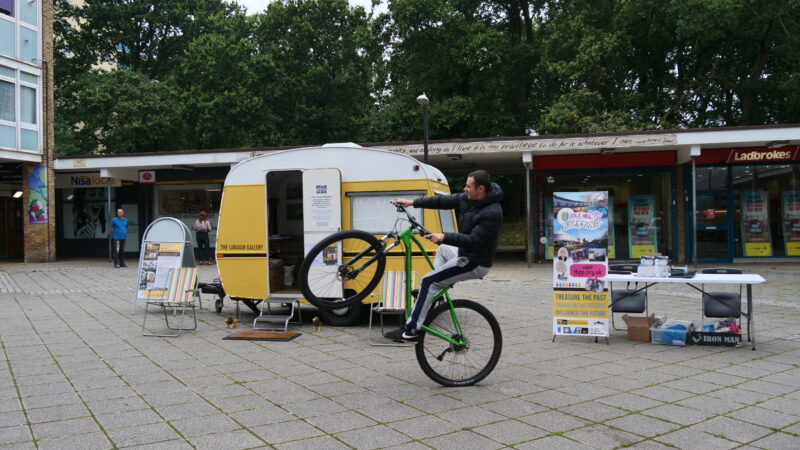 A yellow caravan is parked in front of a row of shops. Two stripey deck chairs are positioned to the side of the caravan. A white man performs a 'wheelie' on a BMX bike in front of the caravan