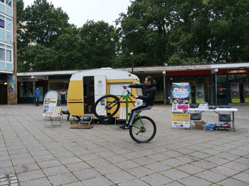 A yellow caravan is parked in front of a row of shops. Two stripey deck chairs are positioned to the side of the caravan. A white man performs a 'wheelie' on a BMX bike in front of the caravan