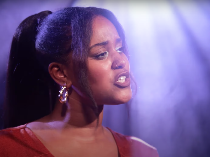 A brown skinned woman with hair in a pony tail sings on stage. The downlighting is dispersed behind her