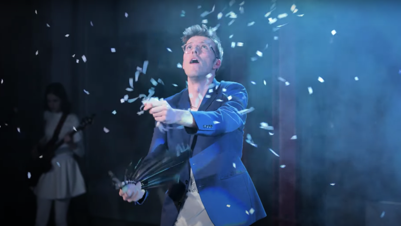 A pale skinned man wears a blue blazer and pale rimmed glasses. He has popped a party popper and confetti flies around him.