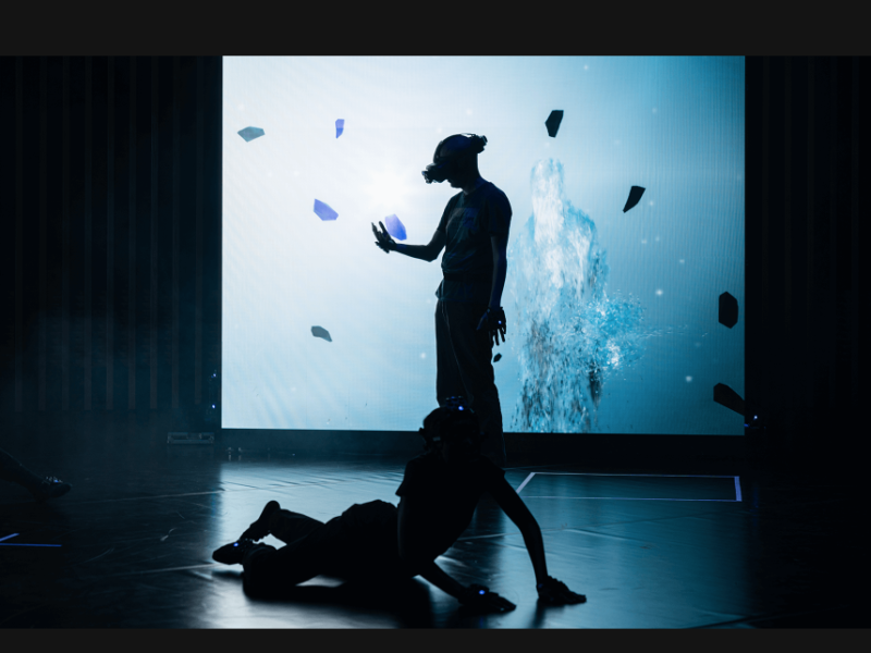 One person stands and is silhouetted against a light blue wall. Another person lies on the floor next to them. The standing person wears a VR headset.