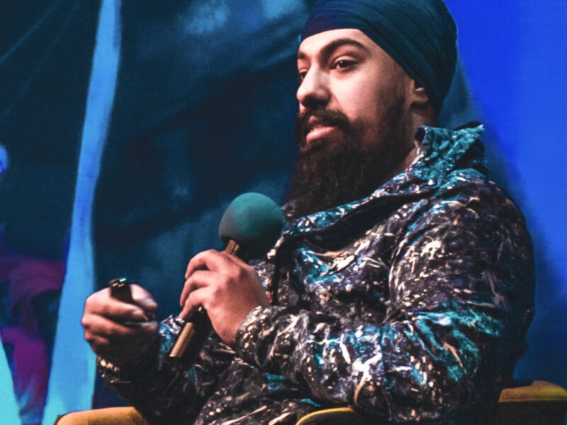 A brown skinned man wears a turban, sitting on stage talking into a microphone