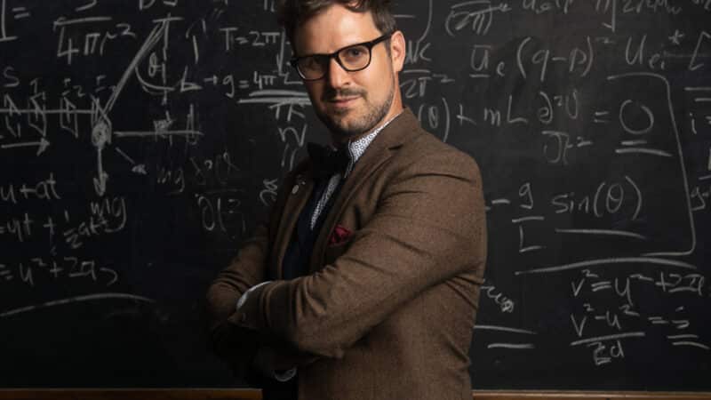 Kevin Quantum dressed in a brown jacket and bowtie with arms crossed in front of a blackboard filled with maths equations