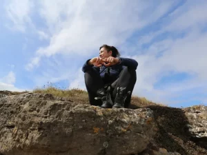 Gina Biggs sitting on a rocky outcrop against a blue cloudy sky.