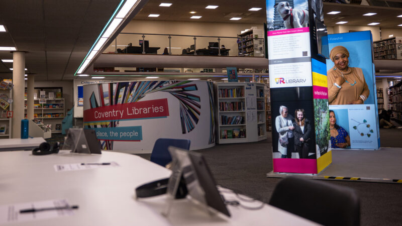 Inside of a library. We see a desk with an ipad and display stands and rows of books in the background