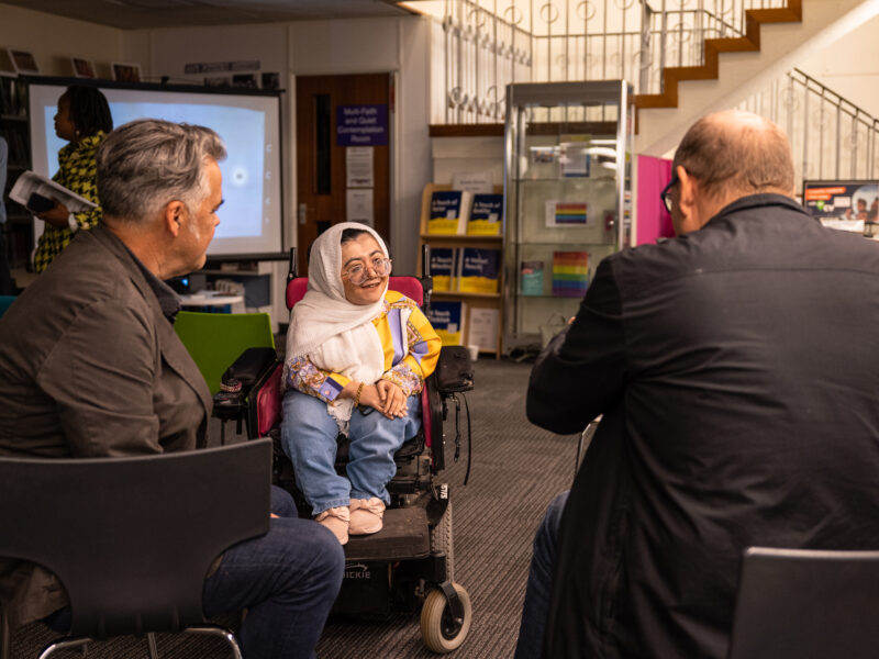 A woman in a wheelchair and a headscarf talks to two seated men