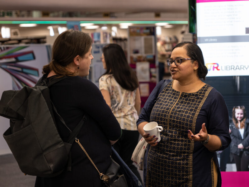Two women, one white, one south asian talk in a library