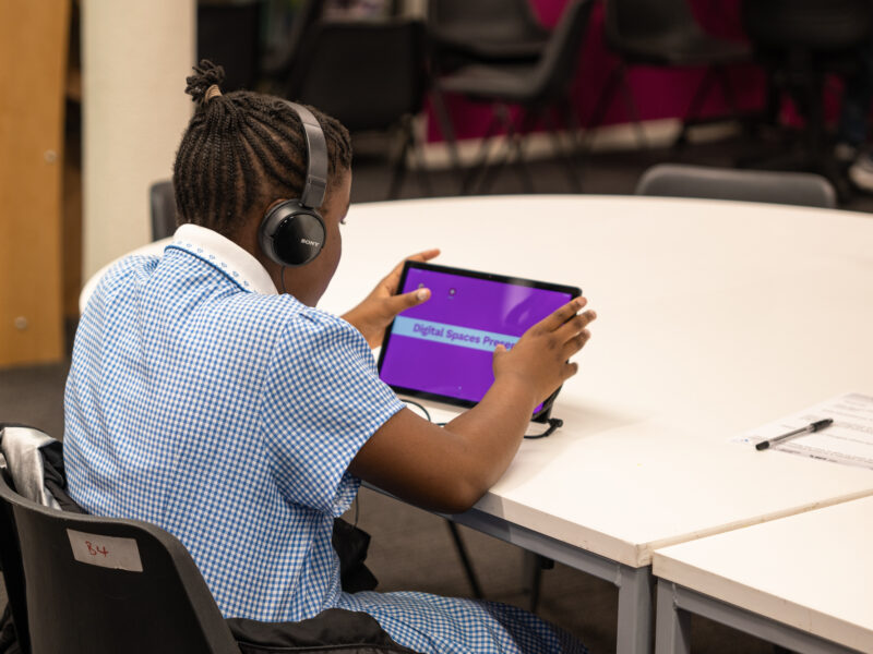 A brown skinned schoolgirl wears headphones and watches a tablet