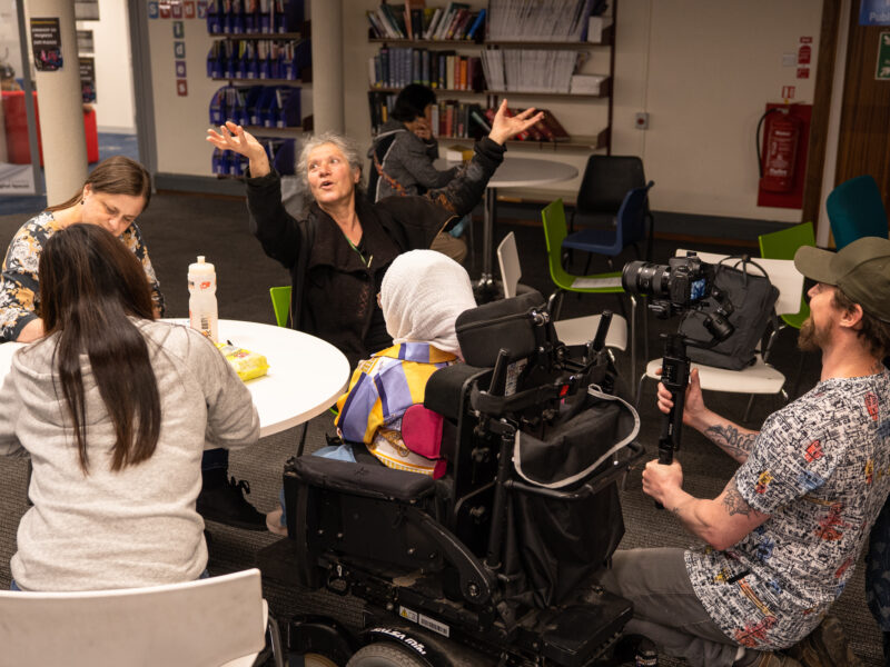 People sit around a table talking. One woman smiles and throws her hands in the air.