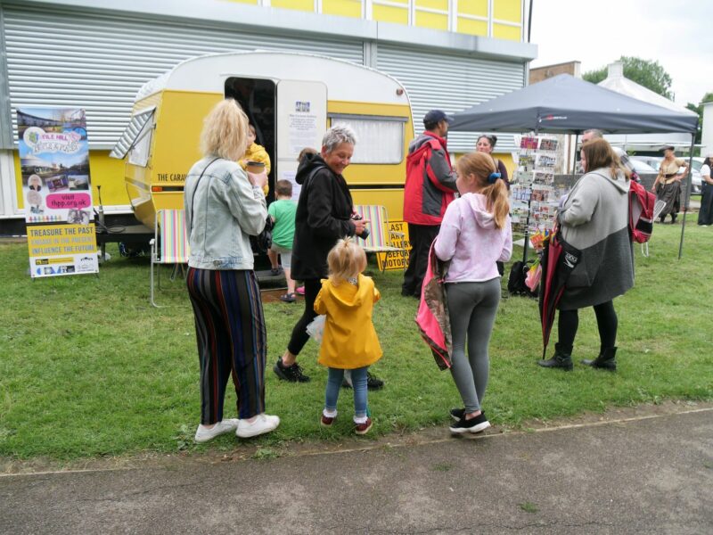 A village gathering, featuring a small yellow caravan and people looking at exhibits and displays