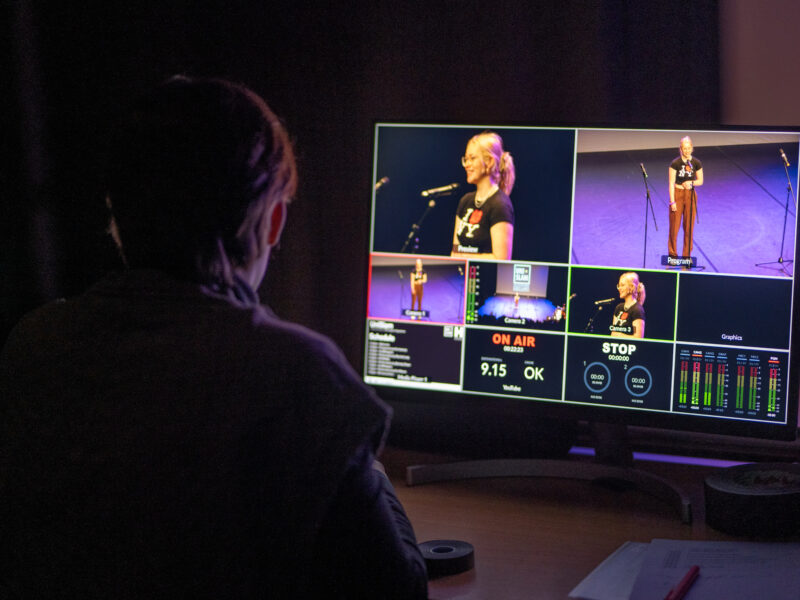 A person sits at an editing screen in the dark. We see images of people performing on stage on the screen