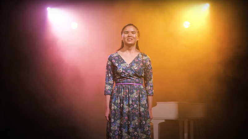 Young woman in a floral dress stands in pink and yellow spotlights reciting poetry