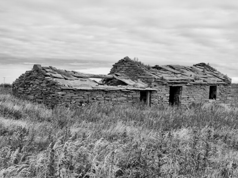 Black and white photo of a dilapidated rural stone brick house surrounded by long grass.