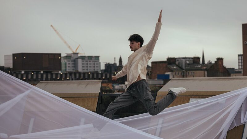 Young male dancer in casual clothes leaping among hung sheets on a rooftop.