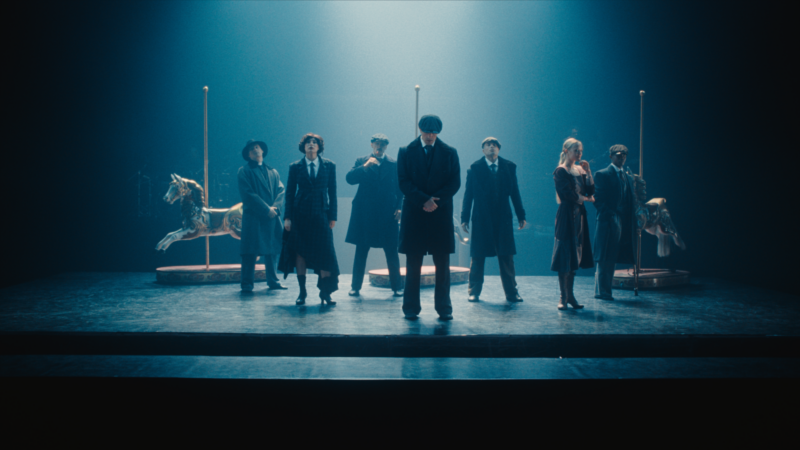 People appear on stage, smartly dressed in 1920's clothing. The lighting is dim and atmospheric. The men wear flat caps. Image from Peaky Blinders: Rambert Dance film