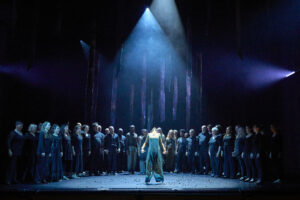 A performer stands at the front of a stage under a spotlight. A long line of other performers appear at the rear of the stage, singing