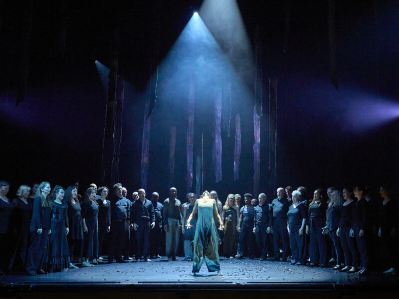 A performer stands at the front of a stage under a spotlight. A long line of other performers appear at the rear of the stage, singing