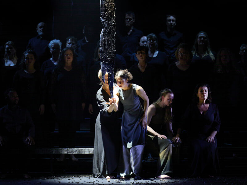 Two performers appear at the front of a stage, one behind the other. They cup their left hands, one under the other. A large shard of metal rests on their hands
