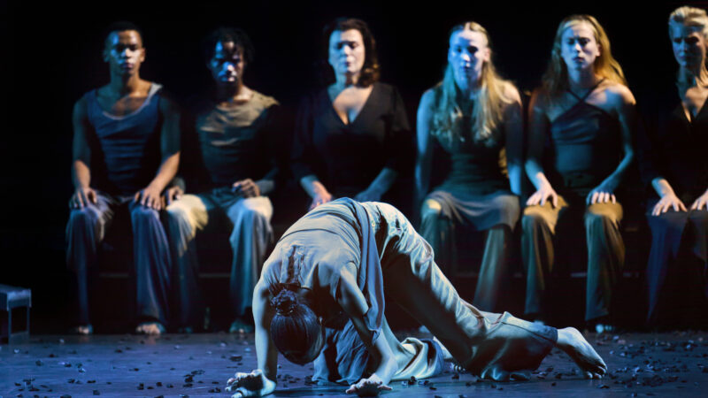A performer is bent double on the floor on stage in front of a row of other performers who are seated, watching.