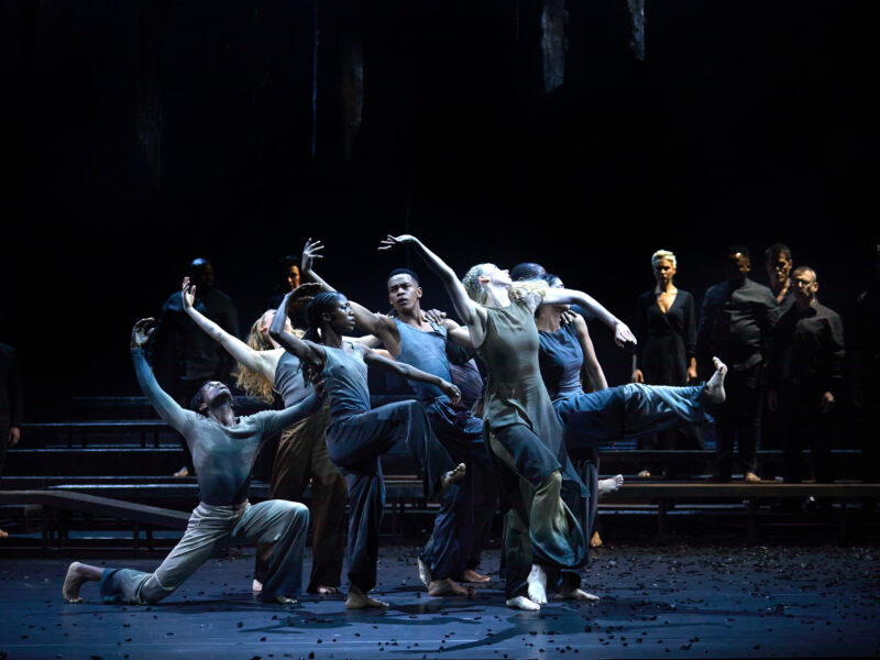 Dancers perform on a dimly lit stage. They appear in an arc and right arms are raised