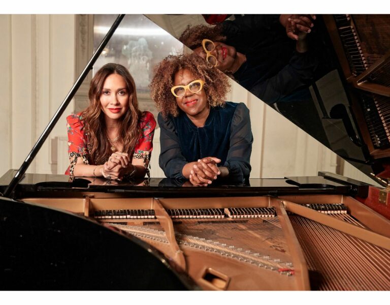 Mylene Klass and Errolyn Wallace rest on a grand piano. The grand piano lid is open