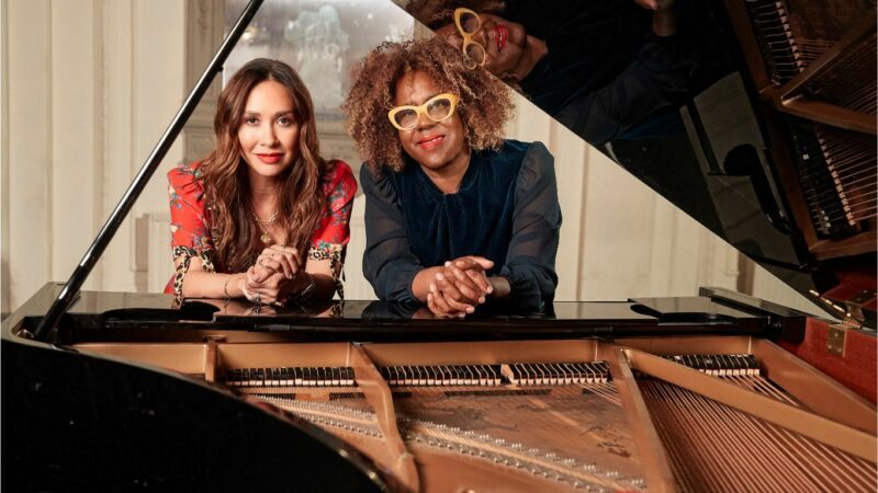 Mylene Klass and Errolyn Wallace rest on a grand piano. The grand piano lid is open