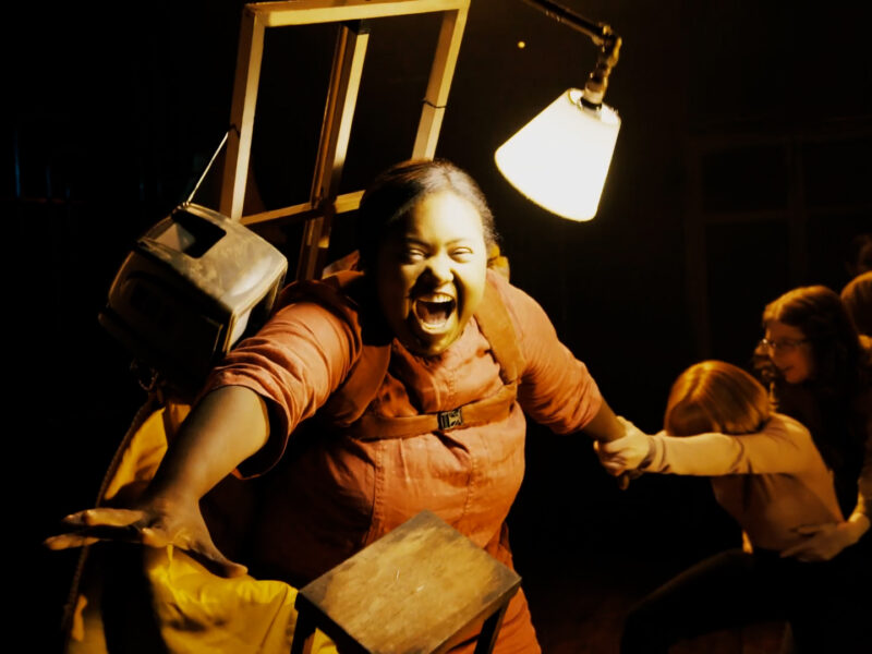 A person performing on a film set. They are illuminated by a lamp and stretch out to their right, whilst being pulled to left by other people.