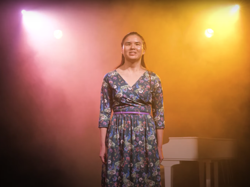A person with visual impairment stands on stage alone. Two bright spotlights shine down upon them. They wear a floral dress and have long dark hair.