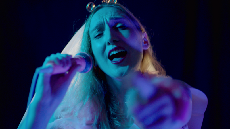 A person wears a bridal headdress and sings into a microphone. They have long blond hair.