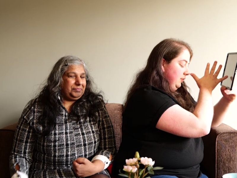 Two people are sat on a sofa. One holds an iPad and is turned away from the other.