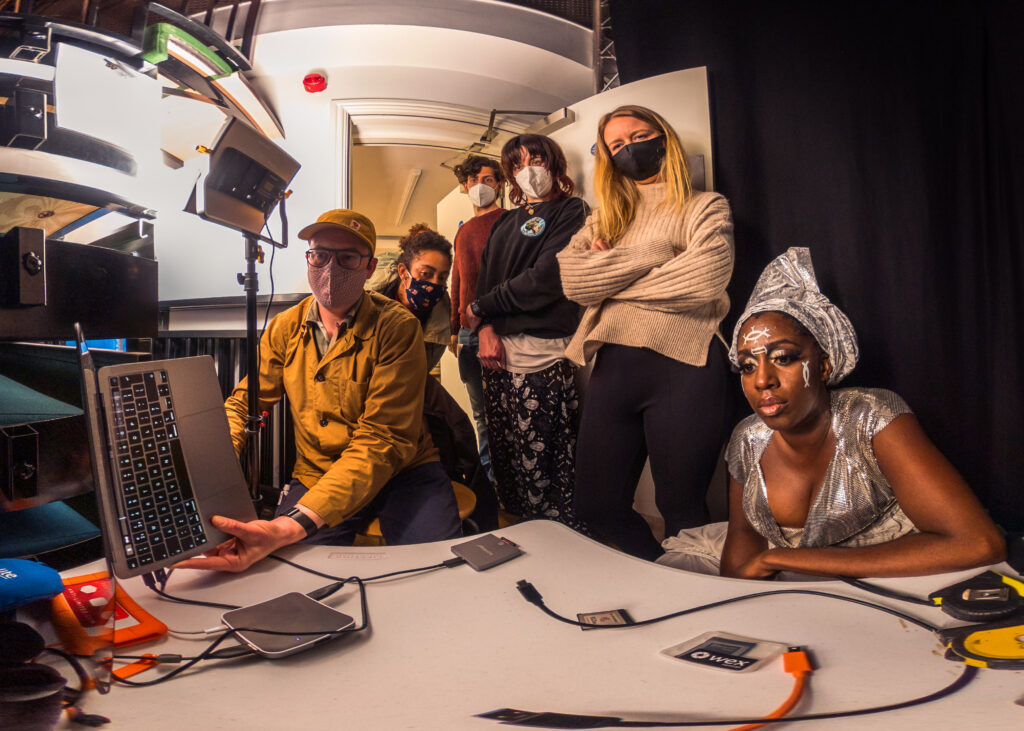 People sit and stand in a small editing studio, all looking intently at a screen. Some wear facemasks.