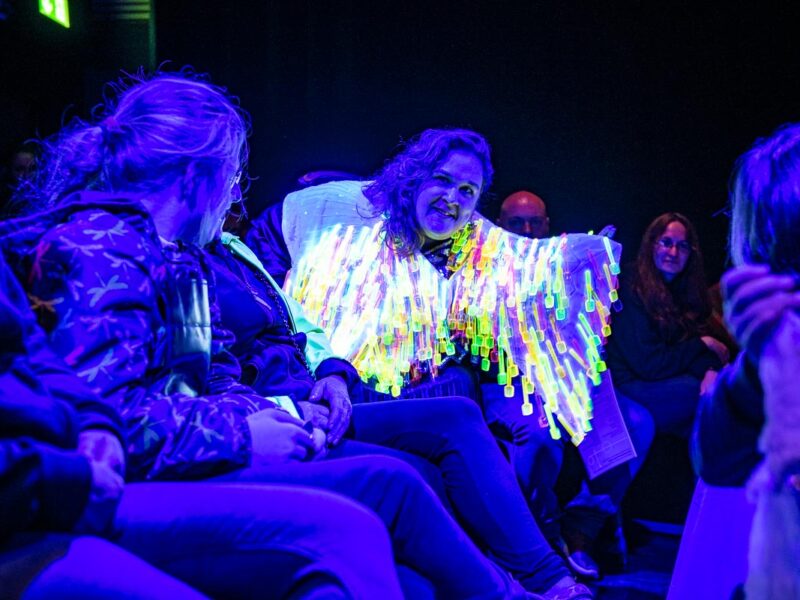 People sit in a line, on chairs. One person wears a 'glow in the dark' jacket, leaning forward.