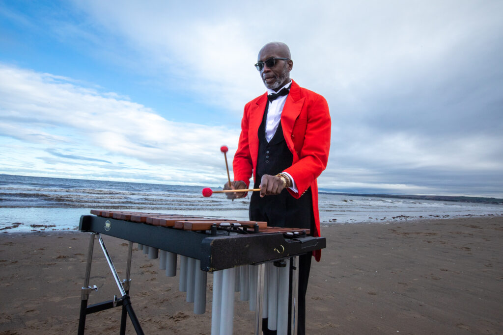 A man in a red tuxedo jacket plays the xylophone on the beach.
