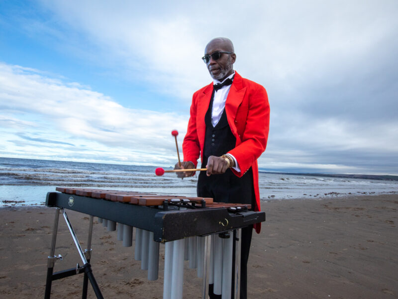 A man in a red tuxedo jacket plays the xylophone on the beach.