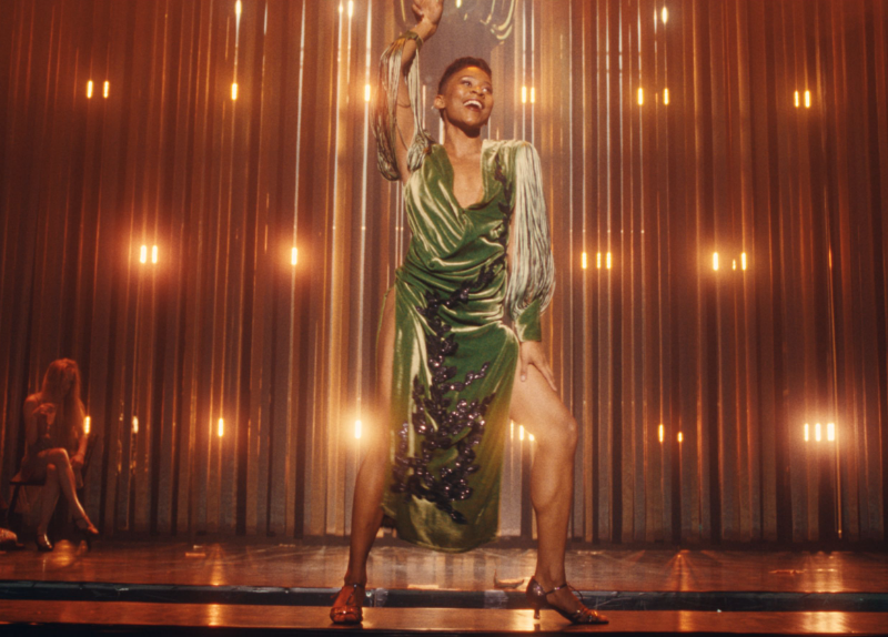 A person with brown skin wears a glamorous green dress and dances on stage
