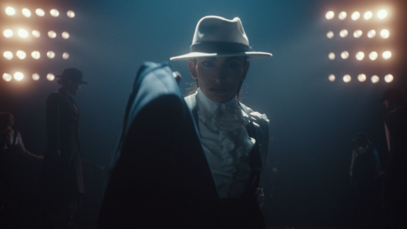 A person is silhouetted by bright links. They wear a white fedora hat and a dark, smart suit