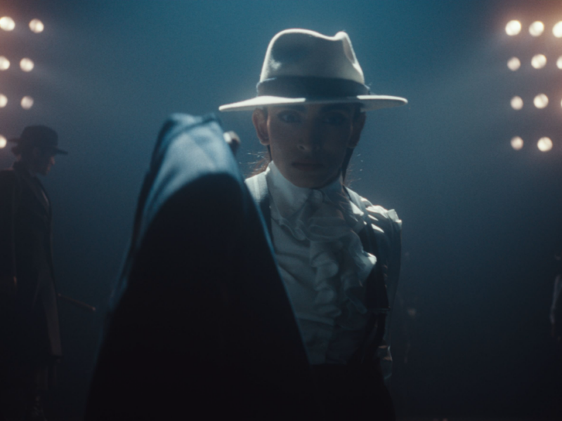 A person is silhouetted by bright links. They wear a white fedora hat and a dark, smart suit