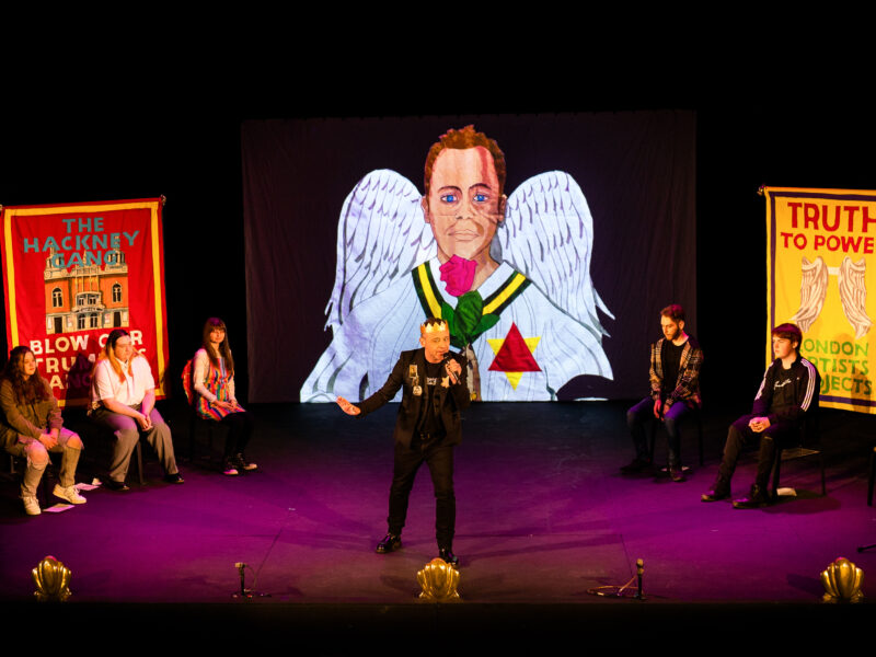A person appears on stage speaking into a microphone. Behind them is a drawing of a cricket player with angel wings.