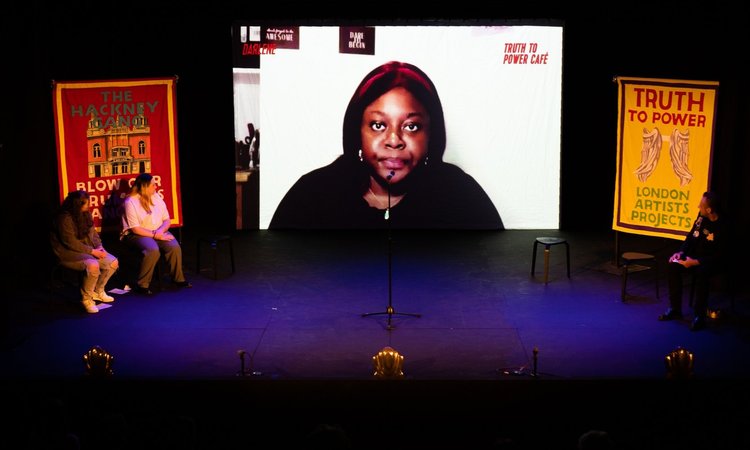 A large screen on stage depicts a person. Flags either side state 'Truth to Power'